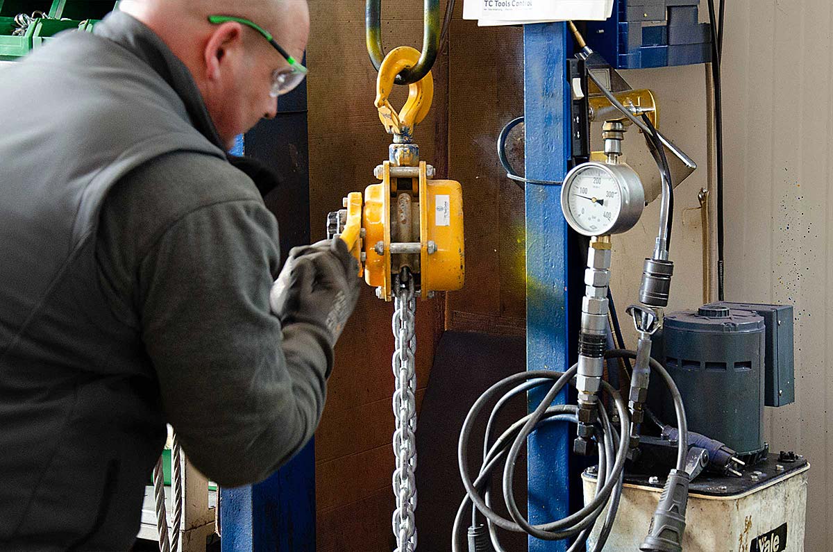 Ein Mann in Schutzbrille und Handschuhen bedient eine gelbe Hebezeugkette in einer Werkstatt mit blauem Metallrahmen und Druckmesser.