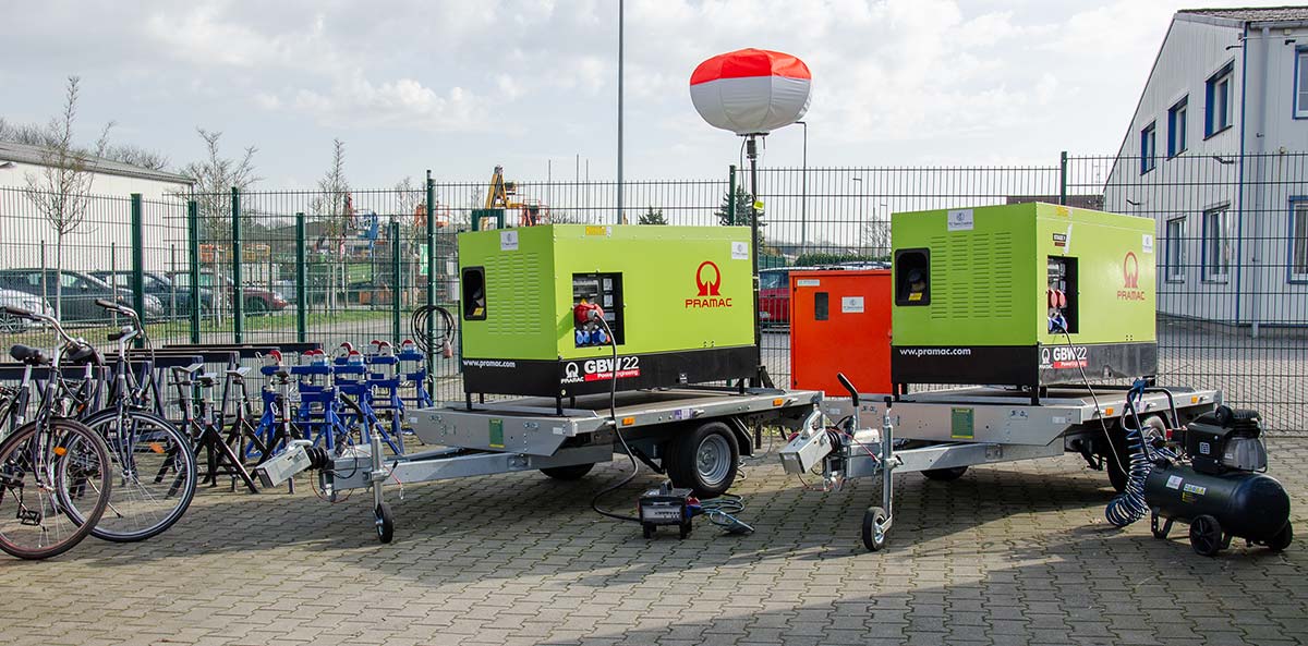 Zwei grüne Pramac-Generatoren auf Anhängern in einem umzäunten Außenbereich, neben Fahrrädern und Baugeräten.