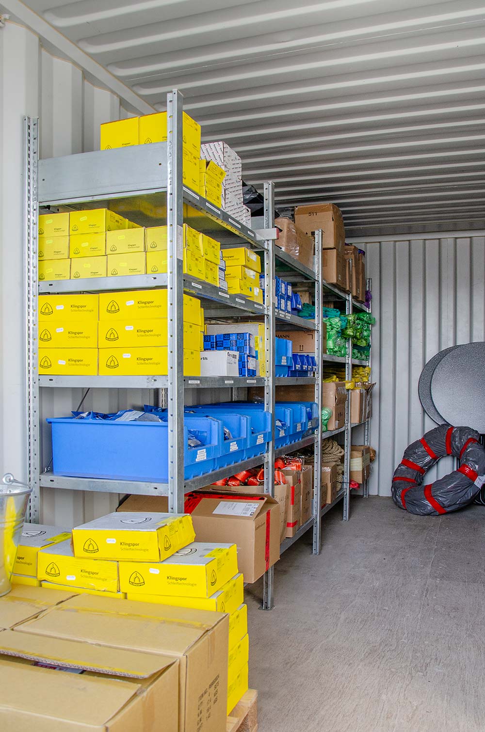 Regale in einem Lagerraum mit gelben Klingspor-Schleiftechnologie-Kartons, blauen Behältern und verschiedenen anderen verpackten Materialien.