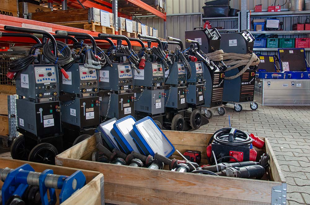 Werkstattausrüstung mit mehreren Schweißgeräten in einer Reihe, Holzboxen mit Abziehwinkelschleifern und Verlängerungskabeln in einer Halle.
