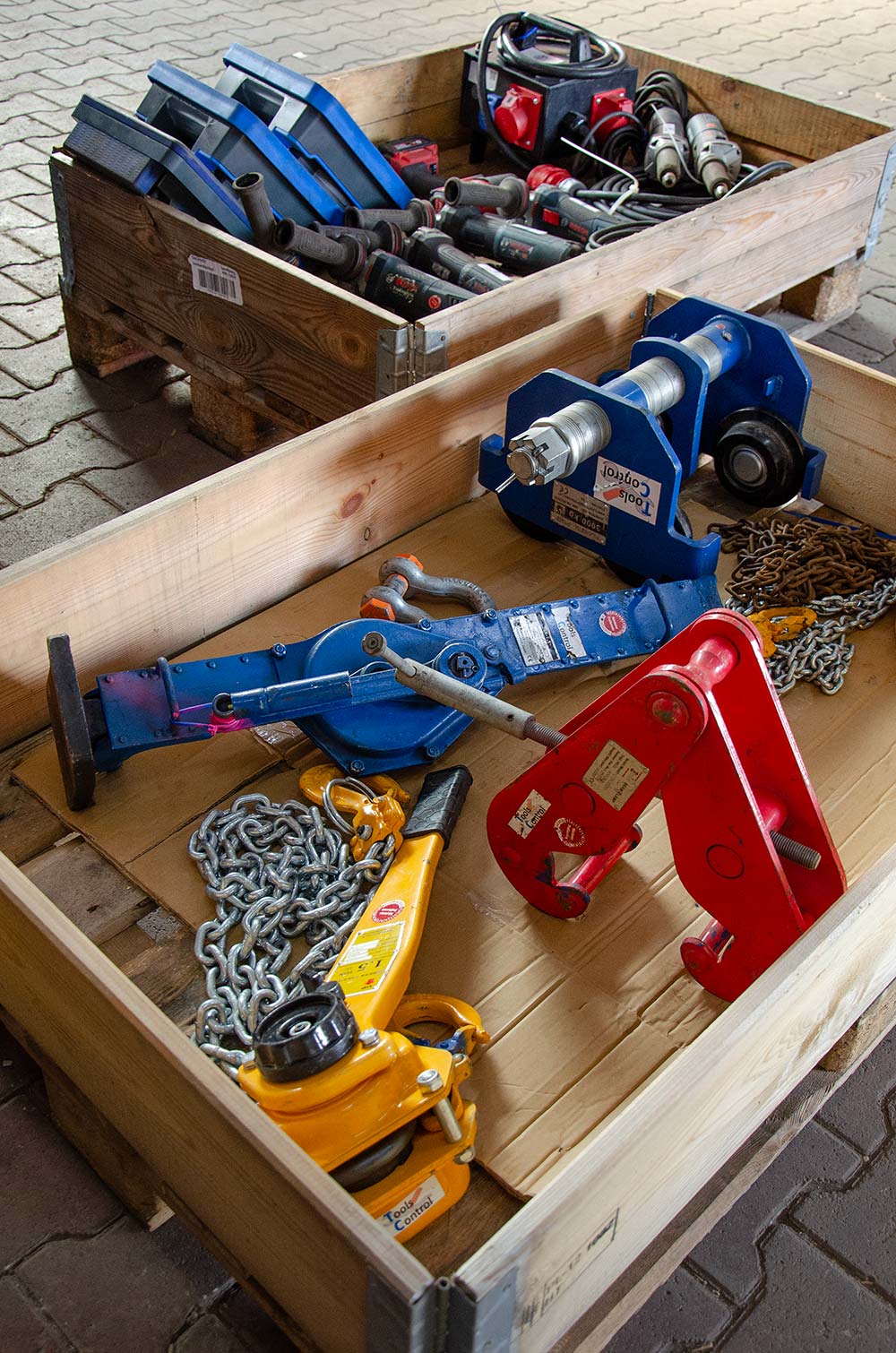 Verschiedene blaue, rote und gelbe Werkzeuge und Kettenteile in zwei hölzernen Kisten auf Pflastersteinen.