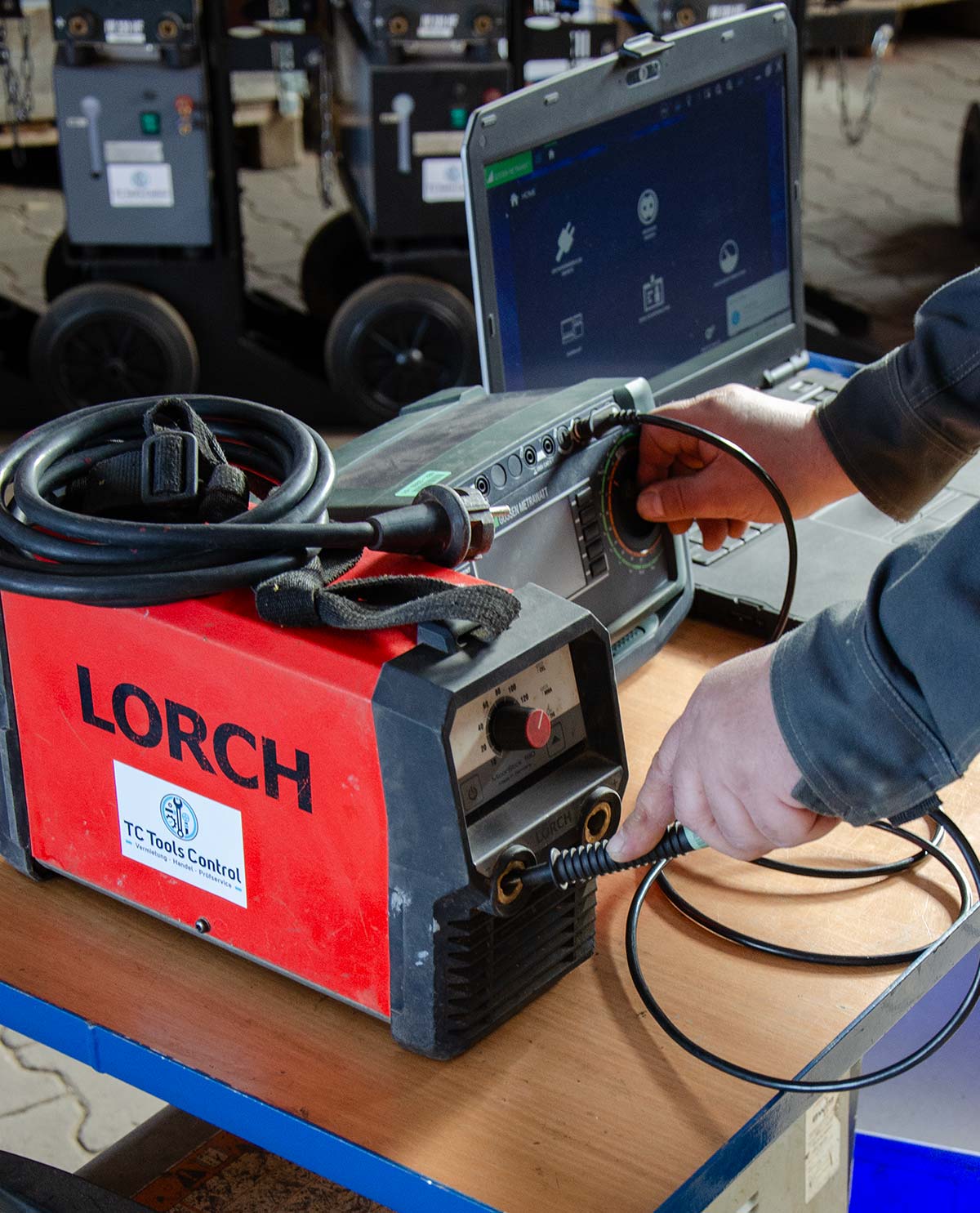 Eine Person verbindet ein Kabel mit einem roten LORCH-Schweißgerät auf einem Holztisch, im Hintergrund ein geöffnetes Laptop mit Kontrolldisplay.