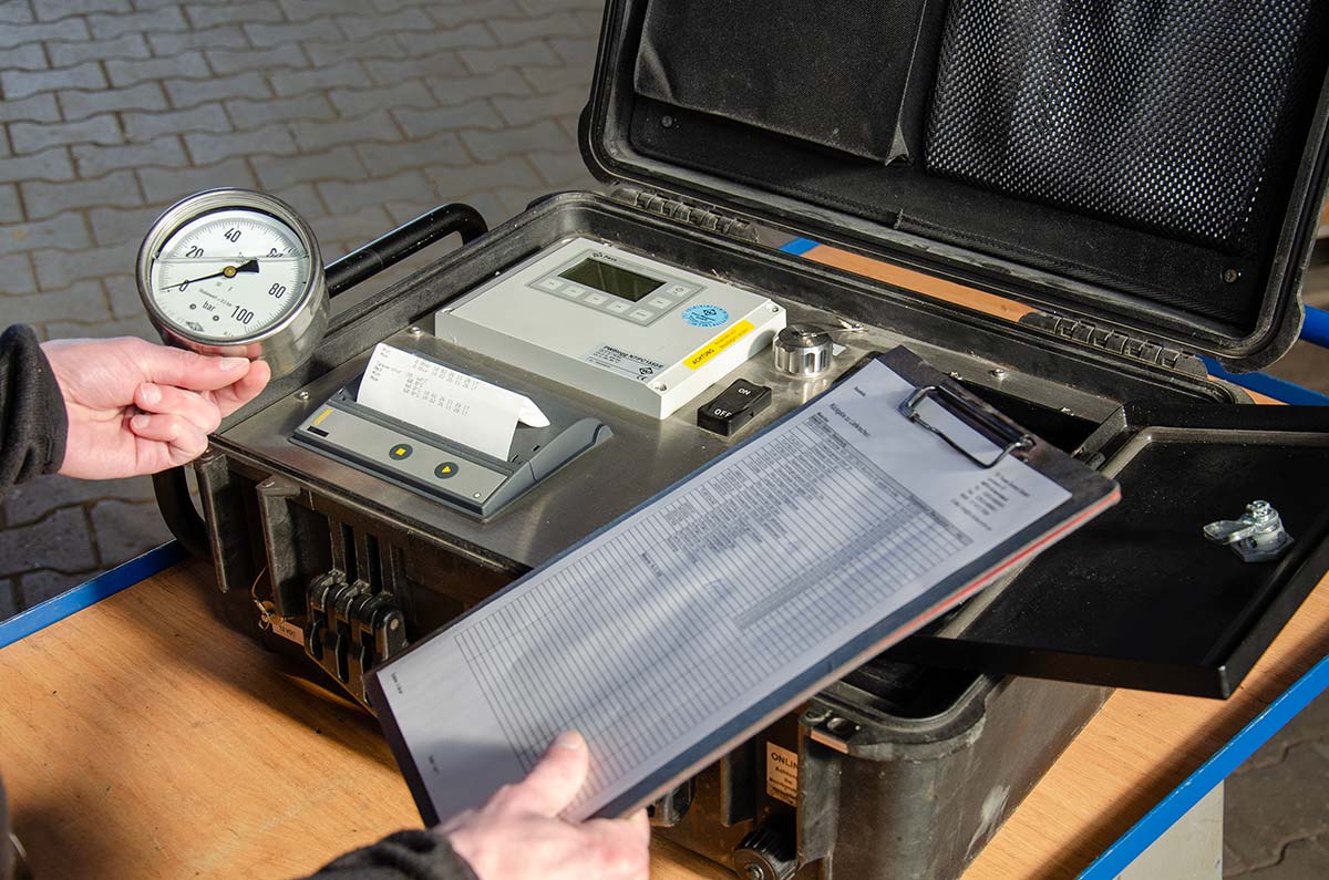 Person hält ein manometrisches Messgerät neben einem geöffneten Koffergehäuse mit einem Druckmessgerät, Druckschalter, eingebautem Thermodrucker und einem Klemmbrett mit einer Tabelle.