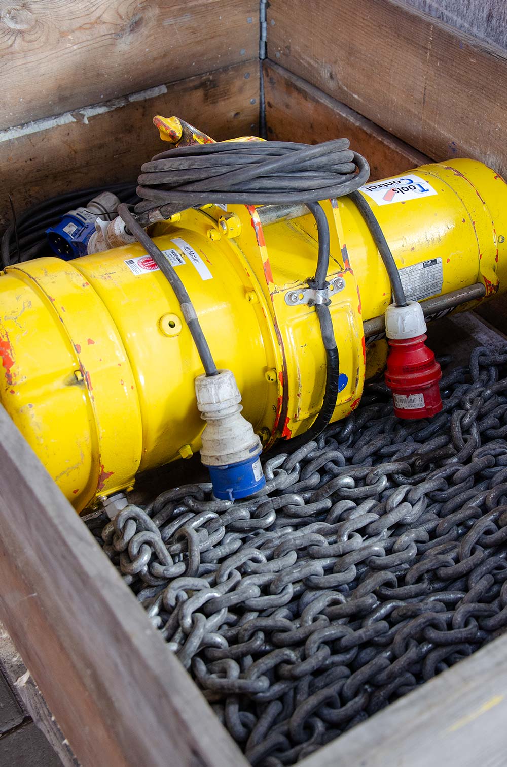 Gelbes elektrisches Werkzeug mit Kabeln, das in einer Holzkiste mit dicken Metallketten liegt.