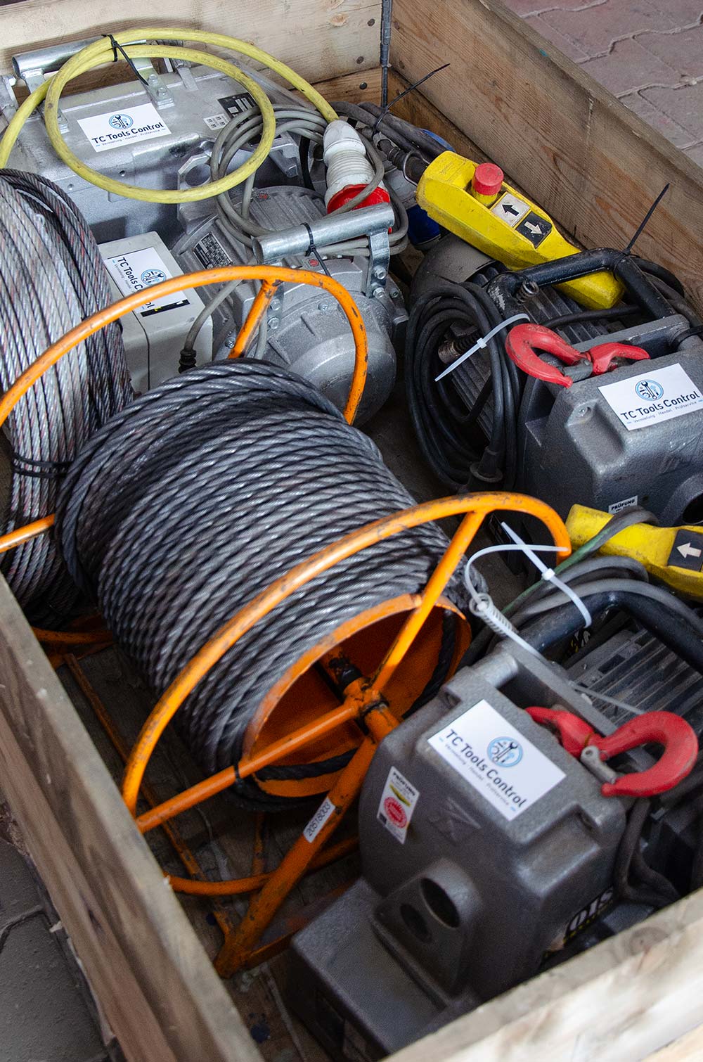 Holzkiste mit zusammengerollter Stahlseilkabeltrommel, mehreren grauen Elektromotoren und gelbem Steuergerät mit roten Not-Aus-Tasten.