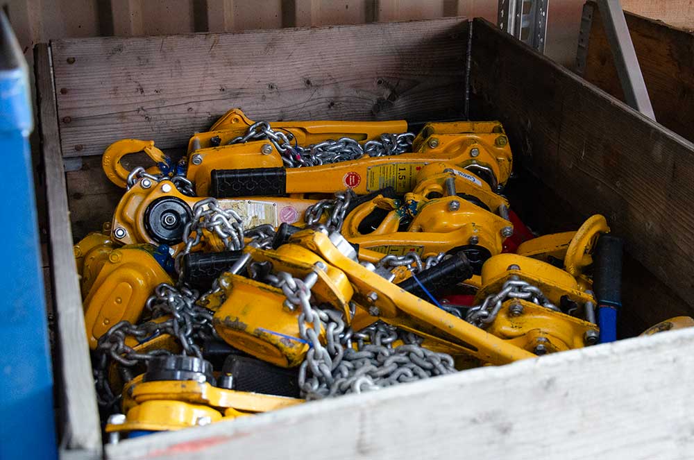 Gelbe Hebelkettenzüge mit Metallketten lagern in einem hölzernen Lagerkasten.